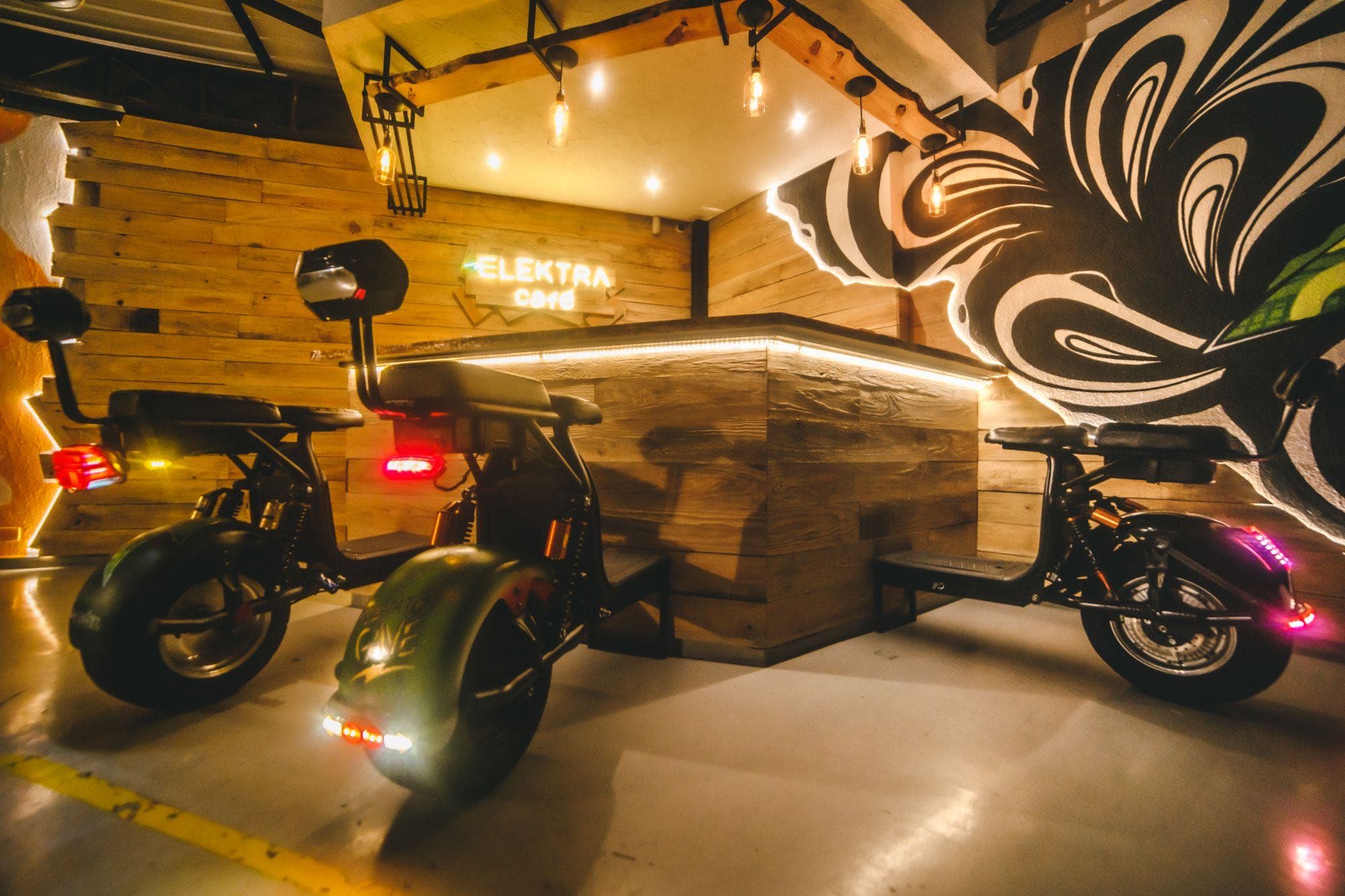 Elektra Café: três scooters elétricos estacionados diante do balcão de madeira iluminado