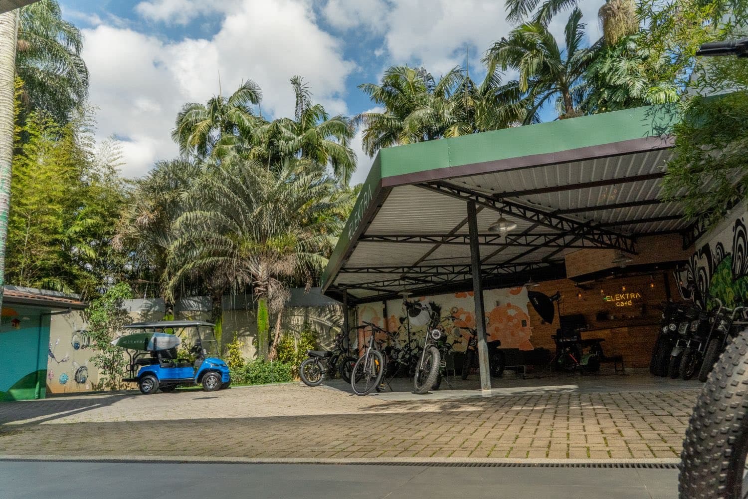 Pátio da Elektra visto de outro ângulo, com bicicletas elétricas sob a cobertura verde e céu azul