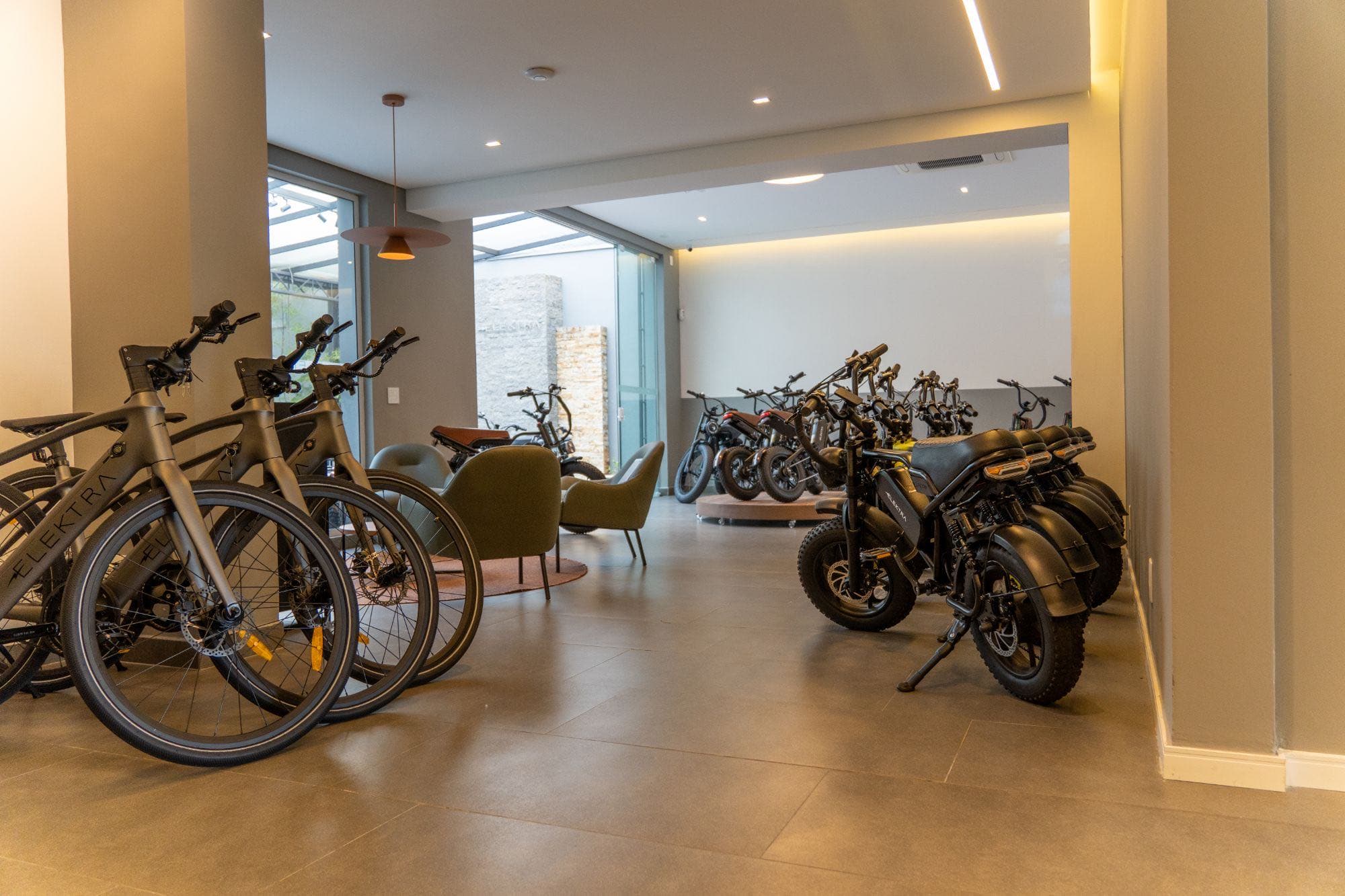 Espaço de espera do showroom, com poltronas verdes, bicicletas e motos elétricas ao fundo