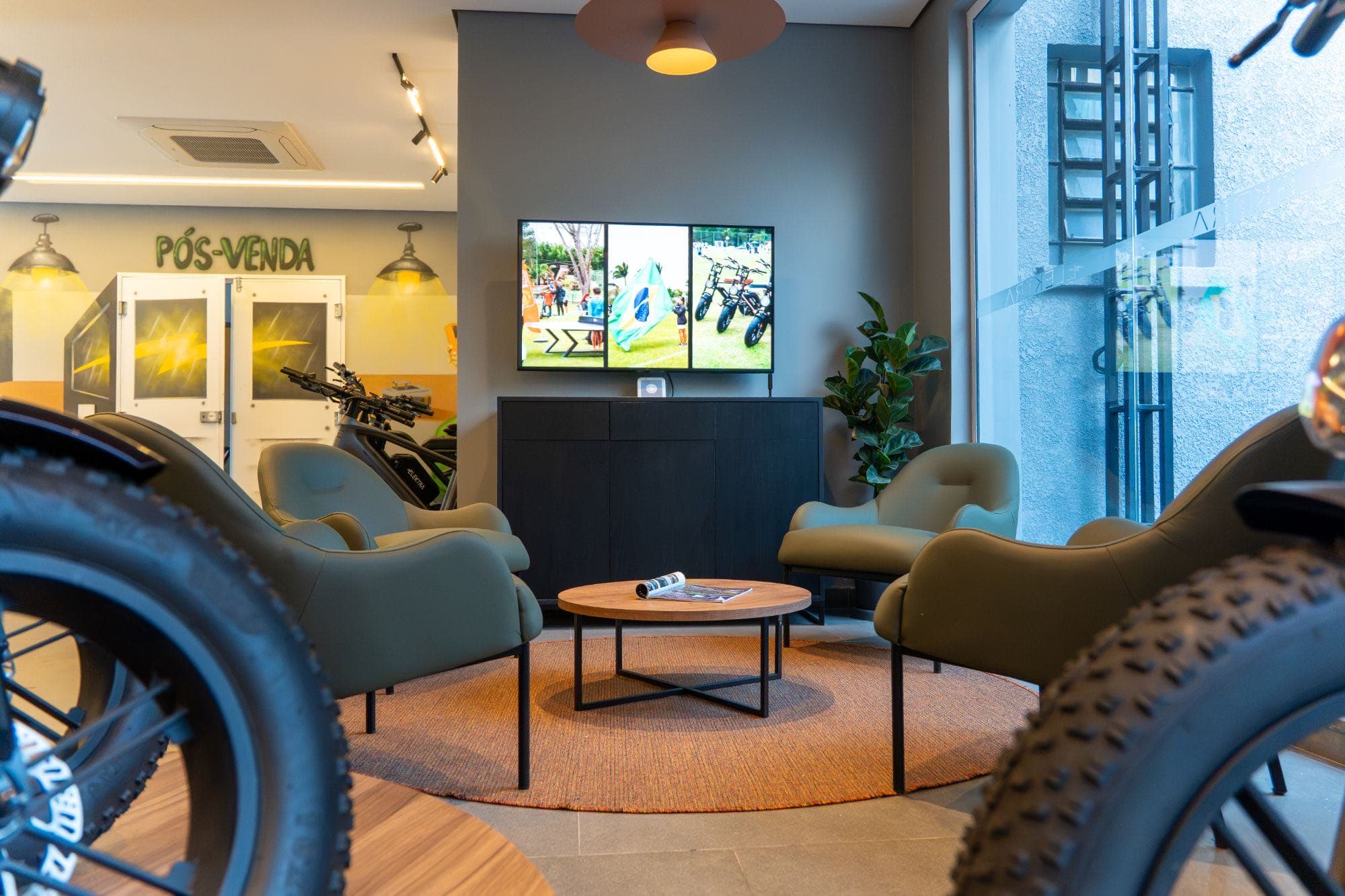 Sala de espera do pós-venda, com poltronas verdes, mesa de centro e TV