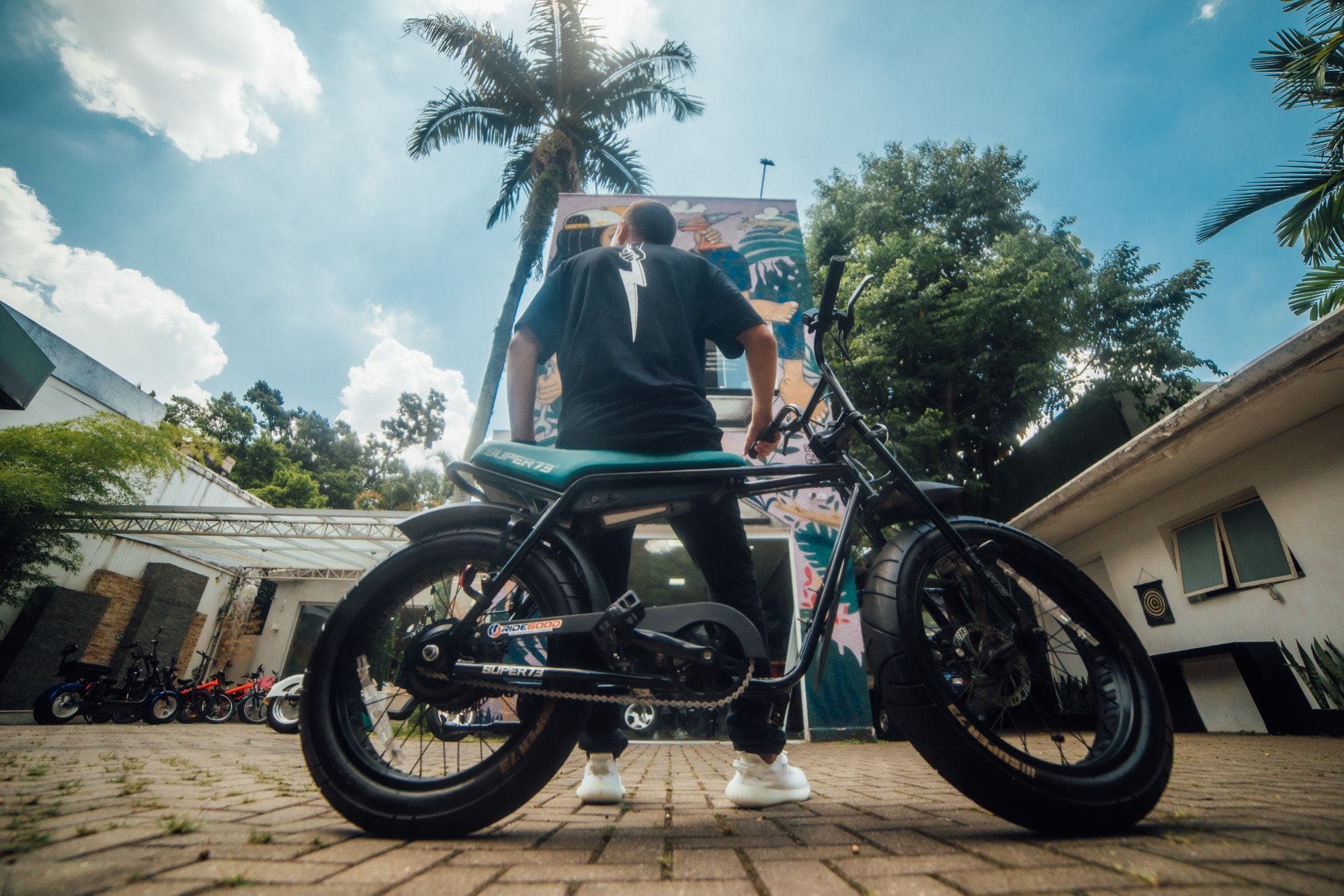 Pessoa de costas sentada em uma bicicleta elétrica no pátio da Elektra, diante da fachada com o mural